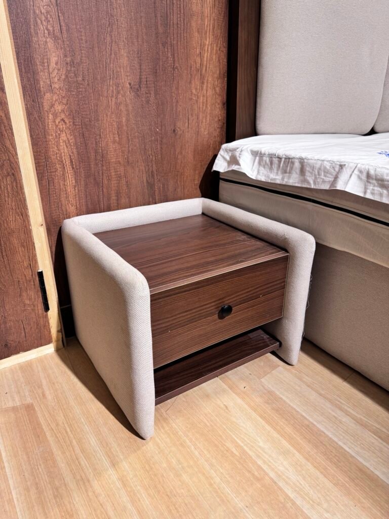 كمود غرفة نوم مودرن بواجهة خشبية بنية ودرج ومقابض سوداء وجانبين منجدين باللون الفاتح، بجوار سرير منجد.